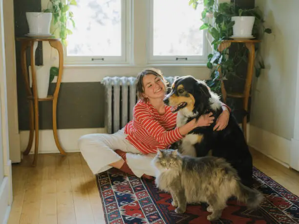 A happy woman sitting on a rug, smiling while hugging her large dog, with a fluffy cat standing nearby in a cozy indoor setting.